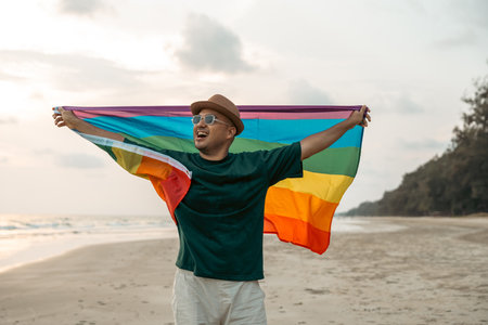 Young asian man with pride movement LGBT holding rainbow flag raise up for freedom. Demonstrate rights LGBTQ celebration pride Month Gay Pride Symbol. Standing on the sand sea beach during sunsetの写真素材
