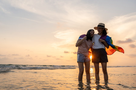 Young couple asian lesbian with pride movement LGBT holding rainbow flag for freedom. Demonstrate rights LGBTQ celebration pride Month lesbian Pride Symbol. Walking on the sand sea beach with sunsetの写真素材