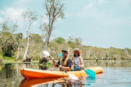 Holiday travel activities. Happy asian couple man and woman rowing a canoe or kayak in mangrove forests. Young traveler with kayak at botanical garden tropical mangrove forest in a national park.の写真素材