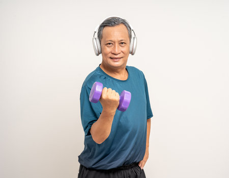 Senior asian old man with sportswear with headset lifting dumbbells on isolated white background. Happy healthy fit and firm attractive sporty mature man standing pose exercise workout in studio.の写真素材
