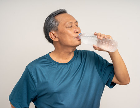 Senior asian old man with sportswear holding bottle of water and drinking on isolated white background. happy healthy fit and firm attractive sporty mature man standing pose exercise workoutの写真素材