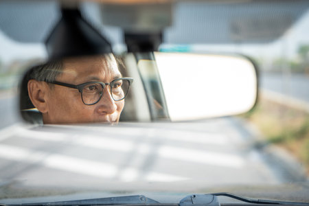 Rear view mirror. Close up shot eye of senior asian man looking roadway while driving car in the morning. Mature Man sitting in car.の写真素材