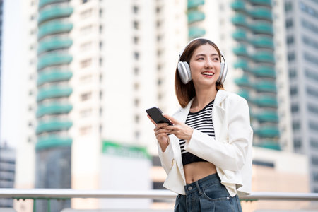 Young asian woman using smartphone while walking in the urban city. Happy Beautiful asian female with headphone listen to the music by cell phone standing at office building city. Lifestyle peopleの写真素材