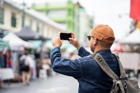 Asian traveling backpacker in Khaosan Road outdoor market in Bangkok Thailand Using mobile phone to take photo. Happy tourist walking in the downtown street traditional marketの写真素材