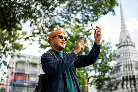 Asian traveling backpacker walking in temple at Bangkok Thailand Using smartphone to take photo and selfie. Tourist walking downtown street traditional ancient temple. Holiday vacation time touristの写真素材