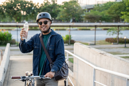 Asian young traveler man riding bike and drinking water. sustainable save energy lifestyle. Commute and carbon neutral transportation. Asian tourist man put on helmet cycling bike to ride urban cityの写真素材