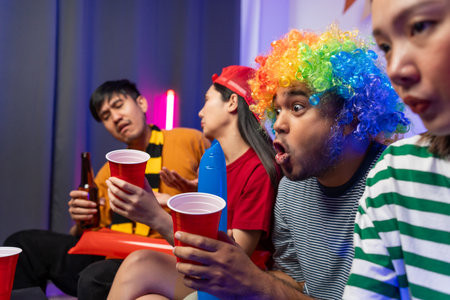 Group of asian friends man and woman watching cheering football match on TV at home. Eating snack and drinking beer. Sport fans people shouting and celebrating sport team victory match.の写真素材