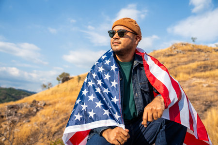 Portrait of cheerful young man looking forward while standing proudly with USA flag over his head against wind and blue sky, happy american holding the United states flag celebrationの写真素材