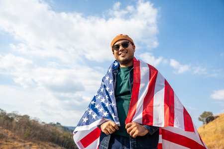Portrait of cheerful young man looking forward while standing proudly with holding USA flag over his head against wind and blue sky, happy american holding the United states flag celebrationの写真素材