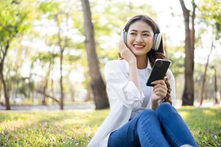 Young asian woman relaxing listening music by smartphone sitting at public park life style outdoor. Happy female Take a break enjoy time living in the nature. Human and nature togetherの写真素材