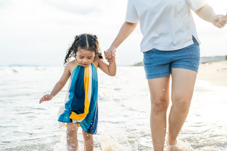 Happy young asian mother and her child walking and holding hands on the beach and enjoy a sunset by the ocean. mom and cheerful kid girl having fun and relaxation on a tropical seaside vacation.の写真素材