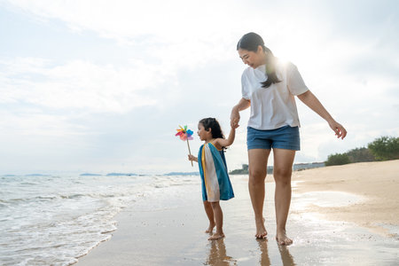 Happy young asian mother and her child walking and holding hands on the beach and enjoy a sunset by the ocean. mom and cheerful kid girl having fun and relaxation on a tropical seaside vacation.の写真素材