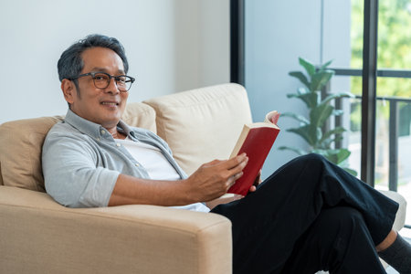 Mature senior Asian old man sitting reading book on cozey couch sofa in living room with sun light morning. People old man stay home part time relax at home reading book.の写真素材