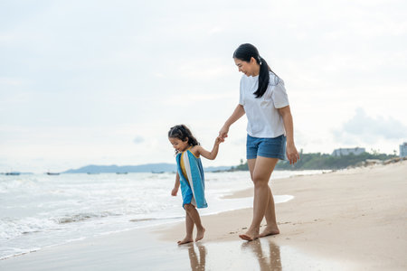 Happy young asian mother and her child walking and holding hands on the beach and enjoy a sunset by the ocean. mom and cheerful kid girl having fun and relaxation on a tropical seaside vacation.の写真素材