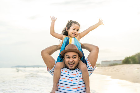 Father carrying his adorable little girl on his back and walking on the beach for family vacation, happy family spend quality time with playing together on summer travel concept.の写真素材