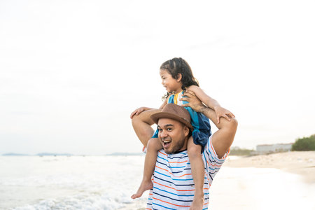 Father carrying his adorable little girl on his back and walking on the beach for family vacation, happy family spend quality time with playing together on summer travel concept.の写真素材