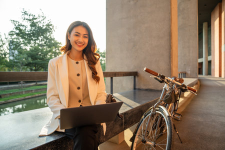 Asian businesswoman riding bike in city working with laptop. Sustainable save energy lifestyle. Commute and carbon neutral transportation. Asian woman working outdoor in the building backgroundの写真素材