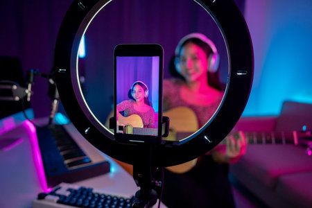 Asian woman musician teaching the guitar class online live streaming with smartphone with student. Female playing acoustic guitar while singing a song and record by cell phone shaed online socialの写真素材