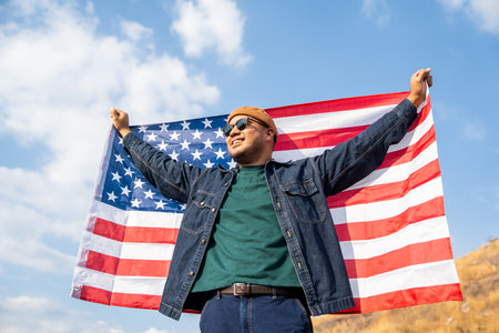Portrait of cheerful young man looking forward while standing proudly with holding USA flag over his head against wind and blue sky, happy american holding the United states flag celebrationの写真素材