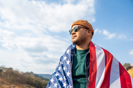 Portrait of cheerful young man looking forward while standing proudly with holding USA flag over his head against wind and blue sky, happy american holding the United states flag celebrationの写真素材