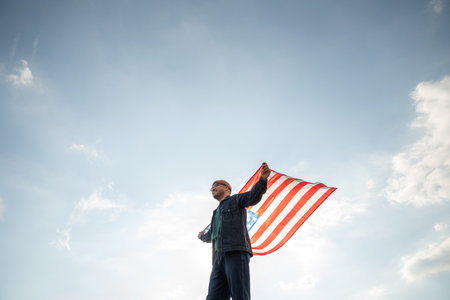 Portrait of cheerful young man looking forward while standing proudly with holding USA flag over his head against wind and blue sky, happy american holding the United states flag celebrationの写真素材