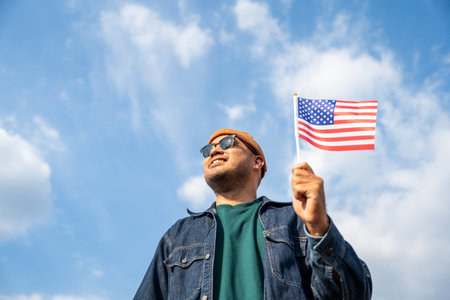 Portrait of cheerful young man looking forward while standing proudly with holding USA flag over his head against wind and blue sky, happy american holding the United states flag celebrationの写真素材