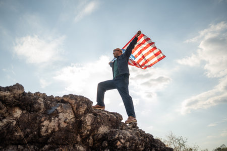 Portrait of cheerful young man looking forward while standing proudly with holding USA flag over his head against wind and blue sky, happy american holding the United states flag celebrationの写真素材