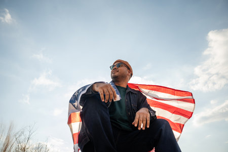 Portrait of cheerful young man looking forward while standing proudly with holding USA flag over his head against wind and blue sky, happy american holding the United states flag celebrationの写真素材