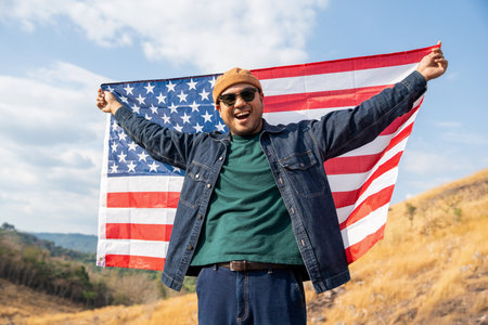 Portrait of cheerful young man looking forward while standing proudly with holding USA flag over his head against wind and blue sky, happy american holding the United states flag celebrationの写真素材