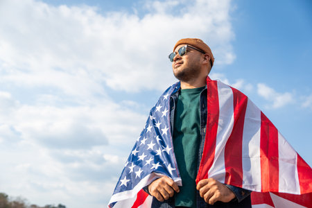 Portrait of cheerful young man looking forward while standing proudly with holding USA flag over his head against wind and blue sky, happy american holding the United states flag celebrationの写真素材