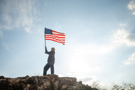 Portrait of cheerful young man looking forward while standing proudly with holding USA flag over his...