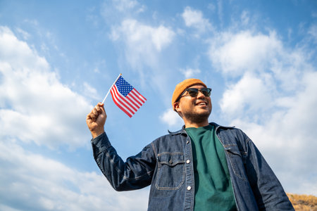 Portrait of cheerful young man looking forward while standing proudly with holding USA flag over his head against wind and blue sky, happy american holding the United states flag celebrationの写真素材