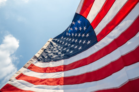 Portrait of cheerful young man looking forward while standing proudly with holding USA flag over his head against wind and blue sky, happy american holding the United states flag celebrationの写真素材