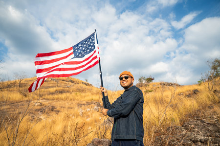 Portrait of cheerful young man looking forward while standing proudly with holding USA flag over his head against wind and blue sky, happy american holding the United states flag celebrationの写真素材