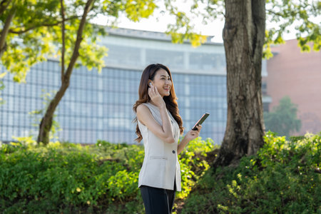 Young asian business woman with headset talking on smartphone standing in the garden public park. Cheerful woman discussion conference on smartphoneの写真素材