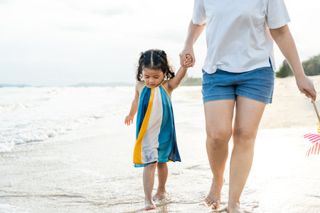 Happy young asian mother and her child walking and holding hands on the beach and enjoy a sunset by the ocean. mom and cheerful kid girl having fun and relaxation on a tropical seaside vacation.の写真素材