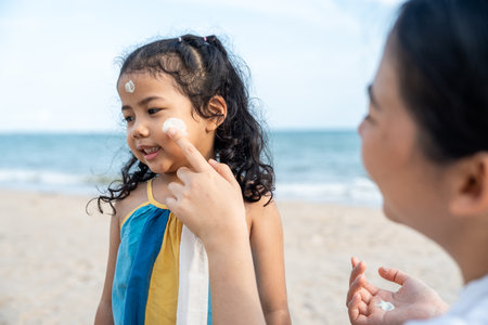 Asian mom applying sunscreen to her happy childâ€™s face on the beach for skin protection from...