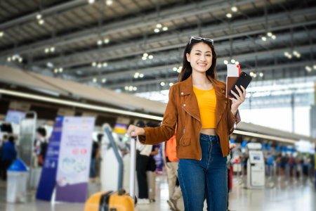 happy young asian woman traveler holding boarding pass ticket, passport and smartphone at the airport terminal with her luggage and smiling at camera, cheerful tourist female having holiday trip.の写真素材