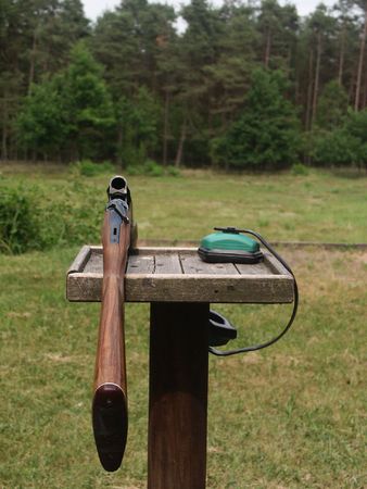Open shotgun and green protective ear coverings sitting unattended on a wooden leaning post. Taken from perspective of the shooter toward a forest area down range.の写真素材