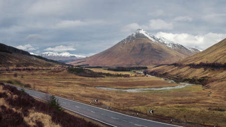 Open road leading through the Scottish Highlands of Glen Coe, snowcapped mountains, and river valleyの写真素材