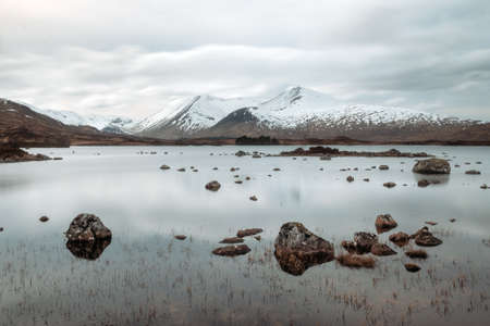 A mountain lake with stones in the water in the foreground. In the background mountains covered with snow. This lake Lochan na h-Achlaise is on the road through Glen Coe, Scotlandの写真素材
