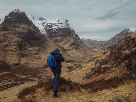 Man photographer with a backpack on the mountain from the back, looking at the mountain valley. The concept of active relaxation, motivation and goal achievement. Glencoe, Scotland, UK.の写真素材