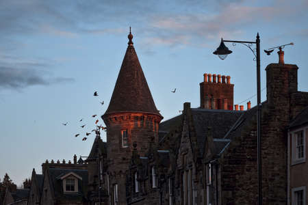 Stone buildings on the street of an old Scottish town with a tower, a lantern and flying birds over the rooftops. Linlithgow, Scotland, United Kingdomの写真素材