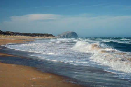 Sea waves on the beach and lighthouse on a cliff. Limetree Walk Beach and Bass Rock, Scotland, United Kingdomの写真素材