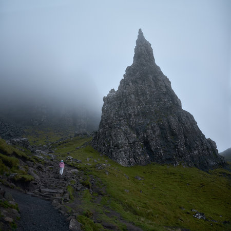 Woman hiker climbs uphill to a high sharp rock covered in fog. The Old Man of Storr covered. The Isle of Skye, Scotland, UKの写真素材