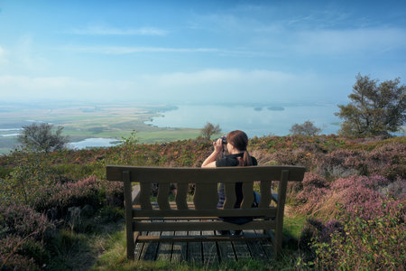 A woman on a bench looks through binoculars in front of a picturesque lake. Vane Hill, Loch Leven, Scotlandの写真素材