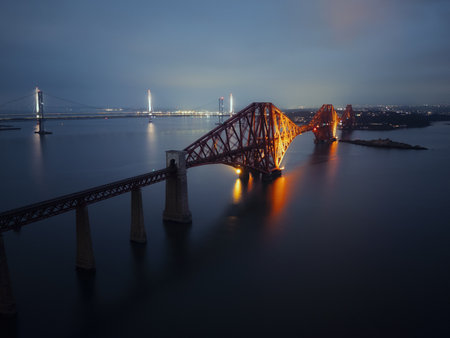 Aerial evening view of the illuminated Forth Bridge in Scotland with reflections on calm water. Iconic landmark captured by drone at dusk.の写真素材