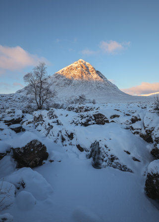 The iconic snow-capped peak of Buachaille Etive Mor in Glencoe, Scotland, bathed in golden morning light. The foreground features a rugged, snow landscape with rocks and a frozen, icy waterfallの写真素材