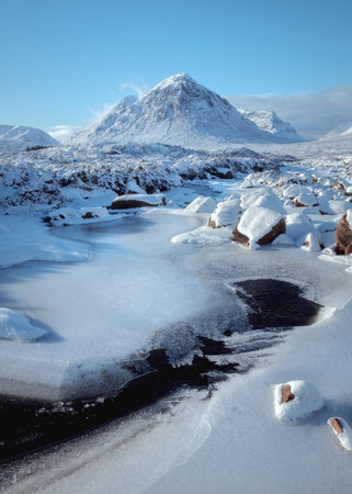 Bright, freezing winter scene in Glencoe featuring the iconic Buachaille Etive Mor rising above the River Etive. The frozen river and snow-covered rocks dominate the foreground under a clear blue skyの写真素材