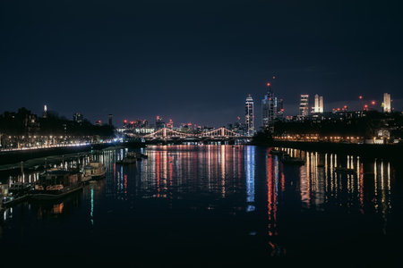 Nighttime view of London with Albert Bridge illuminated over the River Thames. Bright city lights and reflections create a vibrant urban skyline.の写真素材